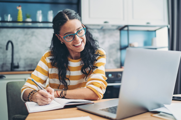 Smiling person taking notes while using a laptop for a virtual Google Meet session in a secure telehealth setting